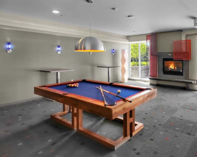 A game room with a pool table, a fireplace, and large windows overlooking a garden area.