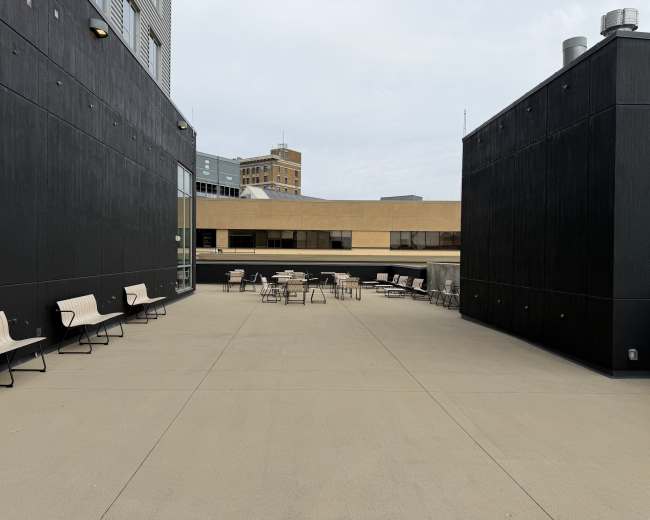 The image shows a rooftop terrace with several tables and chairs surrounded by tall, dark walls and buildings in the background.