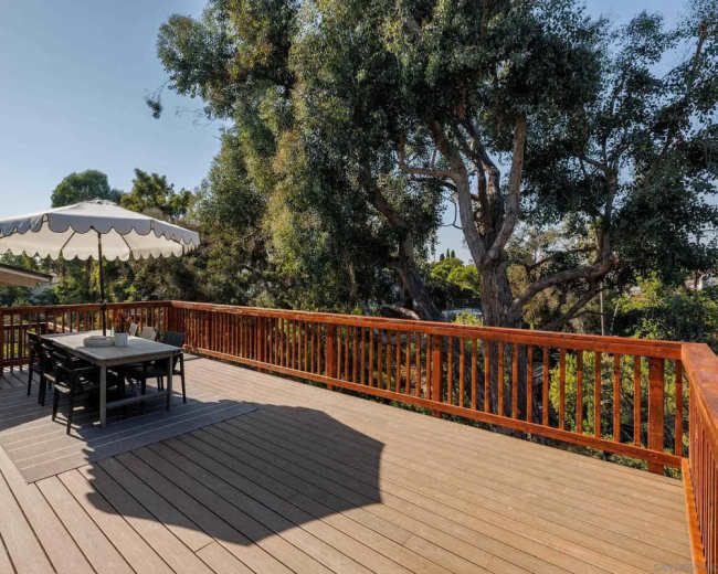 A wooden deck with a dining table and chairs is shaded by a large white umbrella, surrounded by lush greenery.