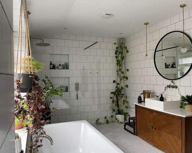 A modern bathroom features a freestanding tub, a large round mirror, plants, and a wooden vanity against a tiled wall.