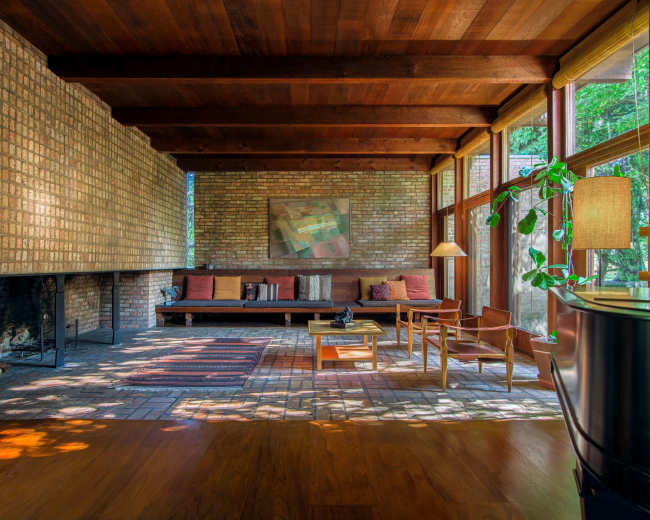 A spacious living room features a brick wall, wooden beams, large windows, and minimal furniture, including a sofa, chairs, and a lamp.