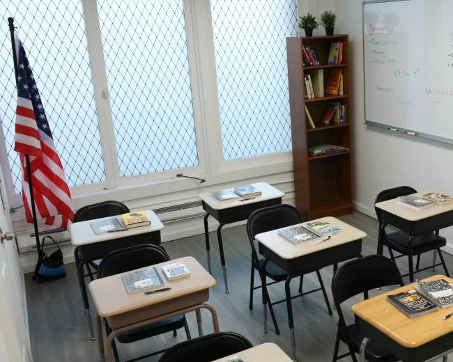 The image shows a classroom with several desks arranged in rows, an American flag in the corner, a bookshelf filled with books, and a whiteboard displaying notes.