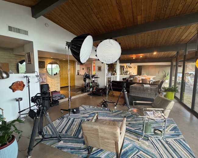 The image shows a well-lit room with a wooden ceiling, equipped with two large light sources, camera equipment, and a patterned rug.
