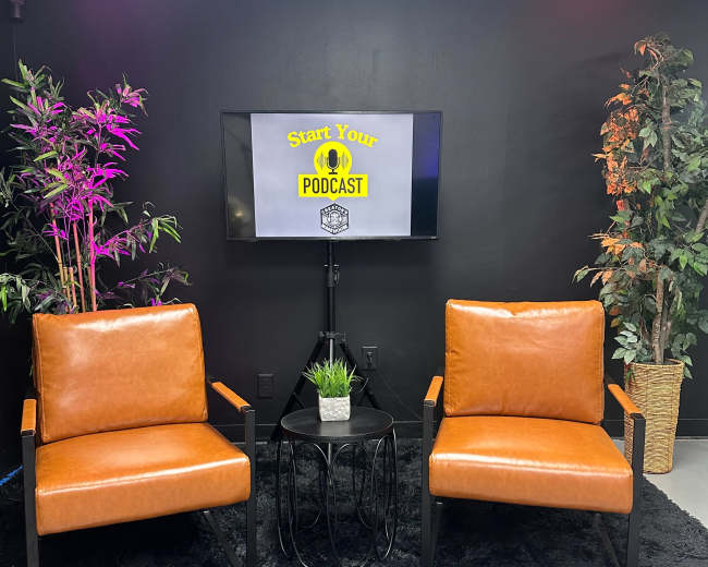 The image shows a podcast setup featuring two brown armchairs, a small black table, and a potted plant against a black wall with a television displaying a sign that reads "Start Your Podcast."