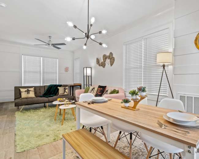 A light-filled living space featuring a dining table with white chairs, a green rug, a sofa with colorful cushions, and decorative wall accents.