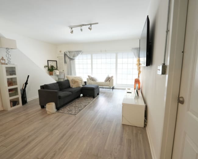 The image shows a well-lit living room furnished with a gray couch, a light-colored armchair, and a TV mounted on the wall, with large windows allowing natural light to fill the space.