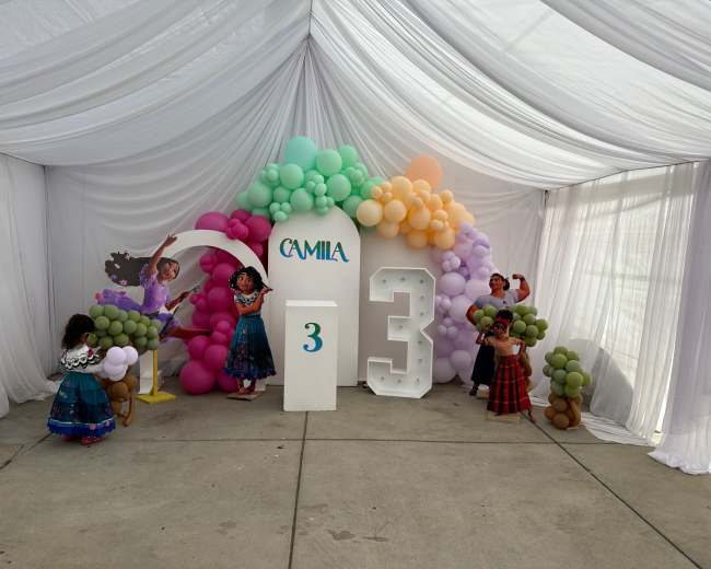 A festive birthday party setup with colorful balloons and decorations, featuring a cake display and the name "Camila" alongside the number "3."
