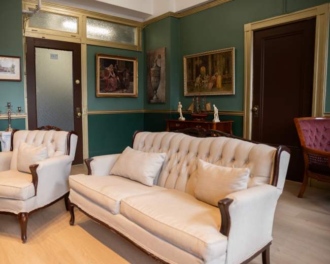 A vintage living room with two upholstered armchairs and a tufted sofa, framed paintings on the walls, and a decorative wooden cabinet.