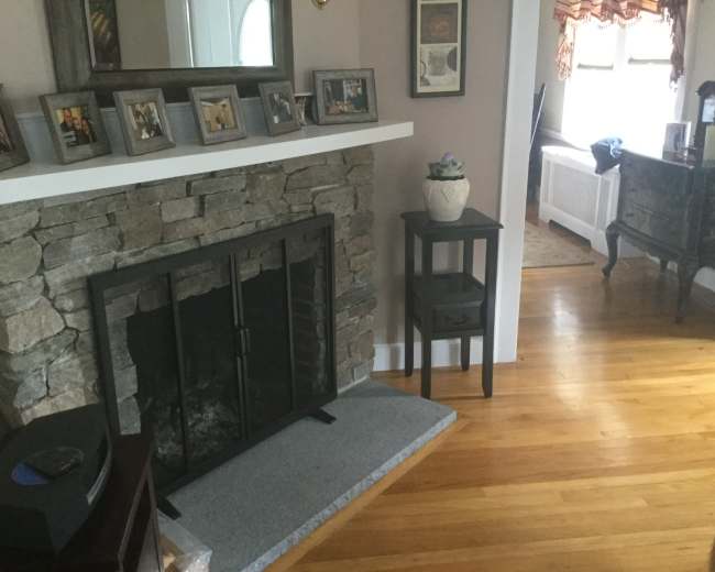 A living room with a stone fireplace, a wooden floor, framed pictures on the mantel, and a small table beside the fireplace.