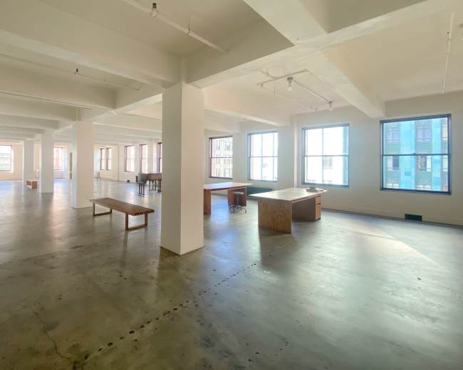 The image shows a spacious, empty office area with large windows and exposed beams.
