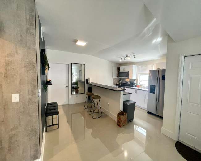 The image shows a modern kitchen with an open layout, featuring a bar-style counter, a refrigerator, and a door leading to another room.