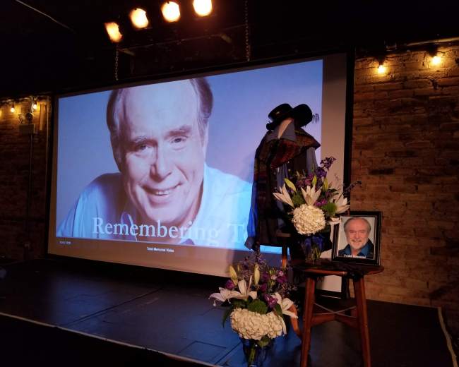 The scene features a stage setup with a large screen displaying "Remembering Them," alongside a framed photograph of a man, flowers, and a hat resting on a microphone stand.