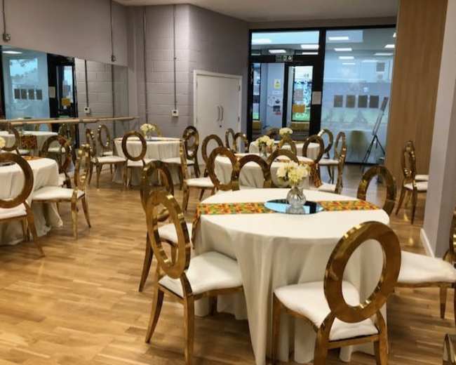 A spacious indoor venue features several round tables with white tablecloths, gold chairs, and floral centerpieces.