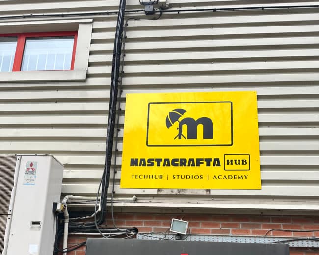 The image shows a building entrance with a large yellow sign displaying the name "Mastacrafta Hub" and its associated labels: "Techhub," "Studios," and "Academy."