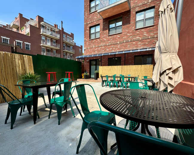 A patio area features green metal chairs and black circular tables, surrounded by brick buildings and a bamboo fence under a clear blue sky.