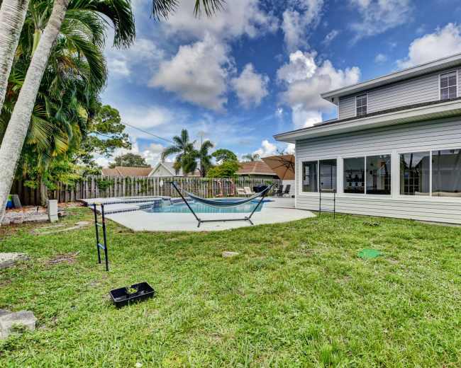 The image shows a two-story house with large windows overlooking a backyard featuring a pool, a hammock, and a grassy area surrounded by palm trees.