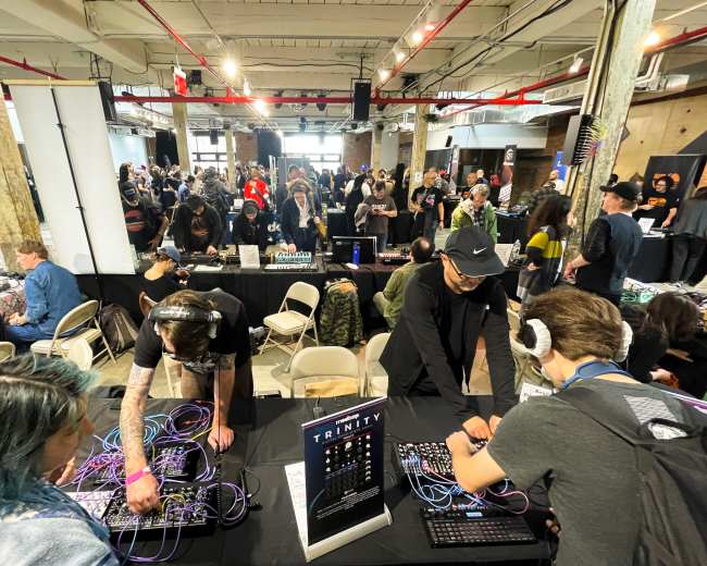 A crowded event space filled with people attending a music or technology festival, with attendees interacting with electronic music equipment at tables in the foreground.