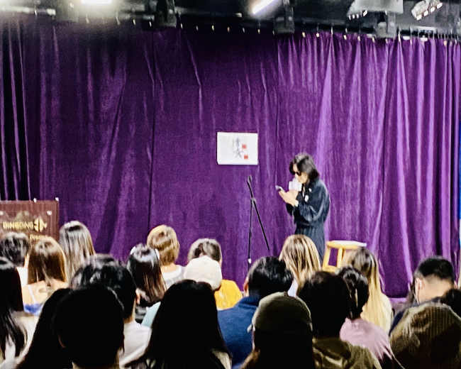 A performer stands near a microphone on stage while an audience sits in front, facing a purple backdrop.