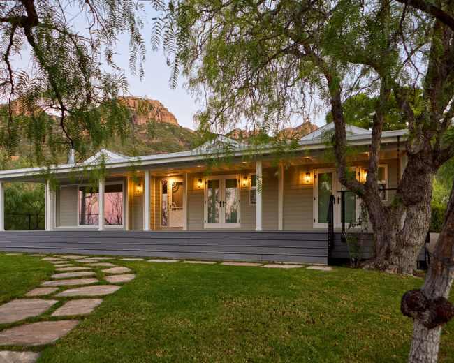 A single-story house with a front porch and large windows is set against a backdrop of mountains, surrounded by green grass and trees.