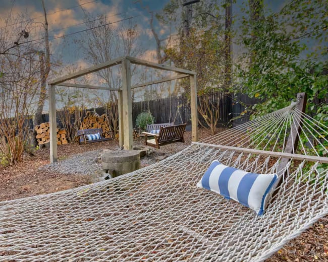 A hammock with a striped pillow is positioned under a wooden frame surrounded by trees and a stone fire pit in a backyard.