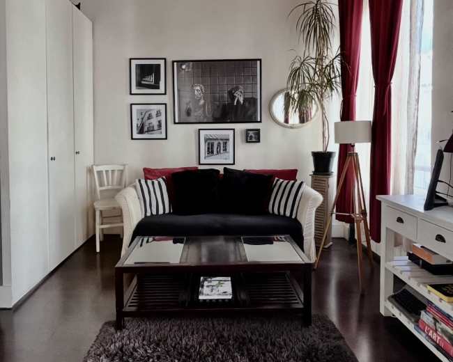 The image shows a cozy living room with a black and white couch, a coffee table, a textured rug, framed photographs on the wall, and large windows draped in red curtains.