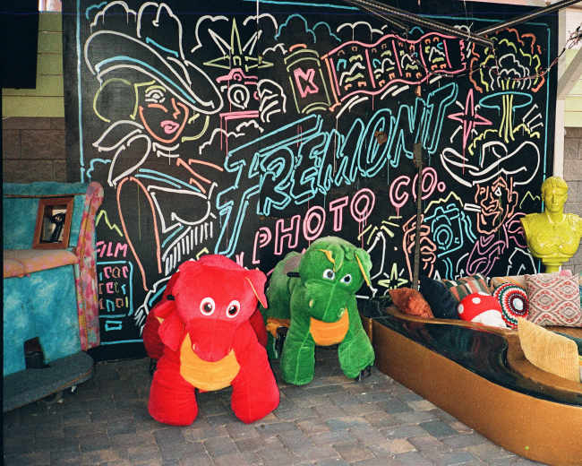 The image shows a colorful wall mural with the words "Fremont Photo Co." and large plush toys in front of it, including a red dinosaur and a green dinosaur, beside a boat-shaped sofa.