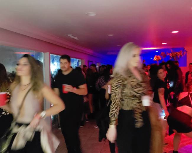 A vibrant party scene features numerous people mingling and dancing under colorful lights, with many attendees holding red cups.