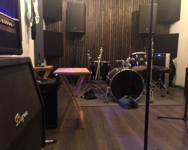 A music practice room with various instruments, including a drum set, amplifiers, and sound equipment, arranged against a wooden wall.