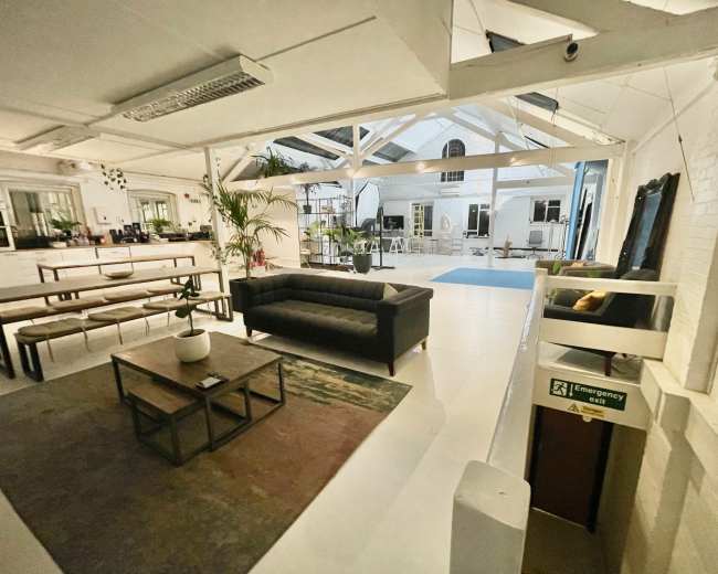 The image shows a spacious, well-lit interior with modern furniture, including a black sofa and a wooden coffee table, surrounded by plants and large windows.