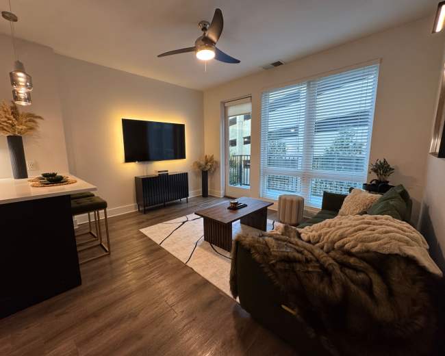 The room features a modern living area with a green sofa, a wooden coffee table, a television mounted on the wall, and large windows that allow natural light to enter.