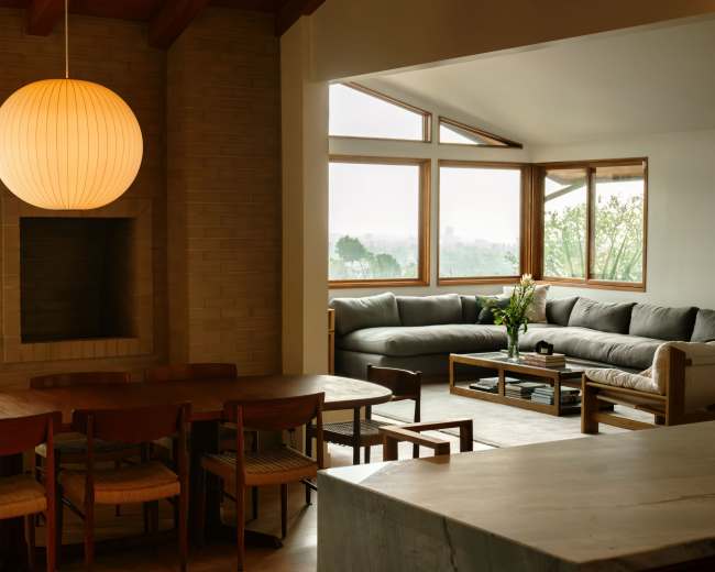 A modern living room features a large gray sectional sofa, a wooden dining table, and a view through large windows.