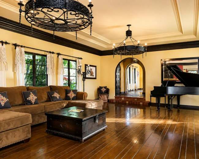 The image shows a spacious living room featuring a large brown sectional sofa, a black grand piano, and two ornate chandeliers, with windows that reveal greenery outside.