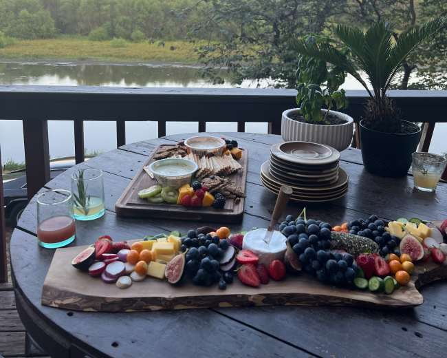 A wooden table displays a variety of fruits, snacks, and drinks, overlooking a scenic river and greenery at sunset.