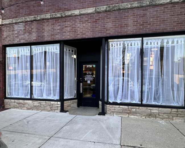 The image shows a brick storefront with large windows dressed in sheer white curtains, revealing some interior lights and decor.