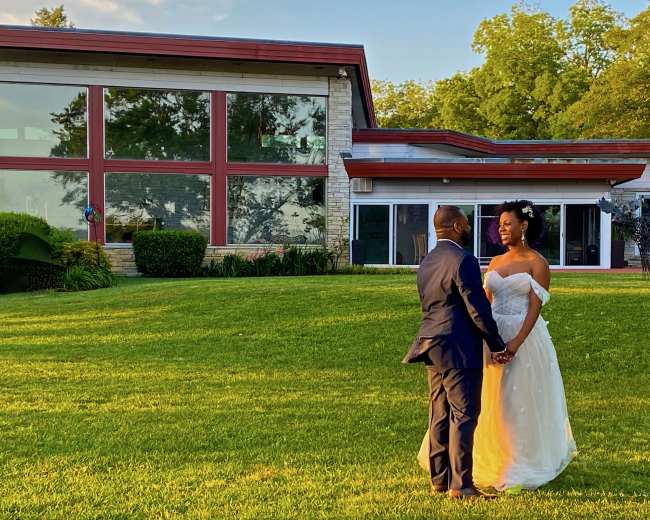 A couple stands holding hands in an open grassy area in front of a modern house with large windows.