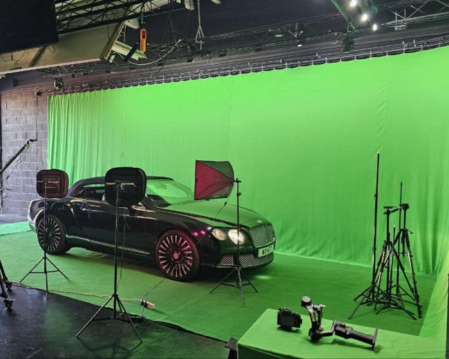 A black luxury car is positioned on a green backdrop in a studio equipped with lighting and camera equipment.