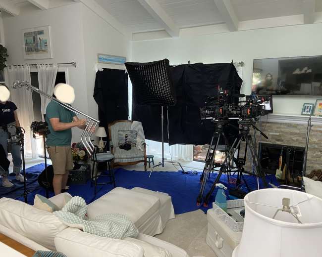A film crew setting up equipment in a brightly lit living room, with cameras, lights, and sound equipment arranged around a seating area.