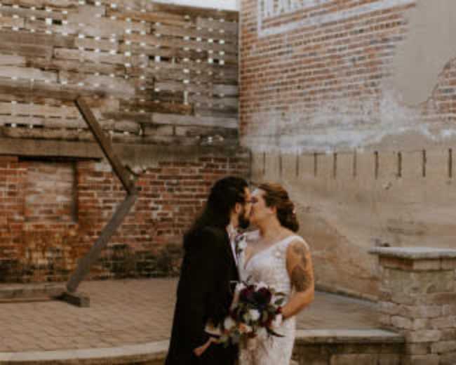 A couple exchanges a kiss in a rustic courtyard with exposed brick walls and a stone structure.
