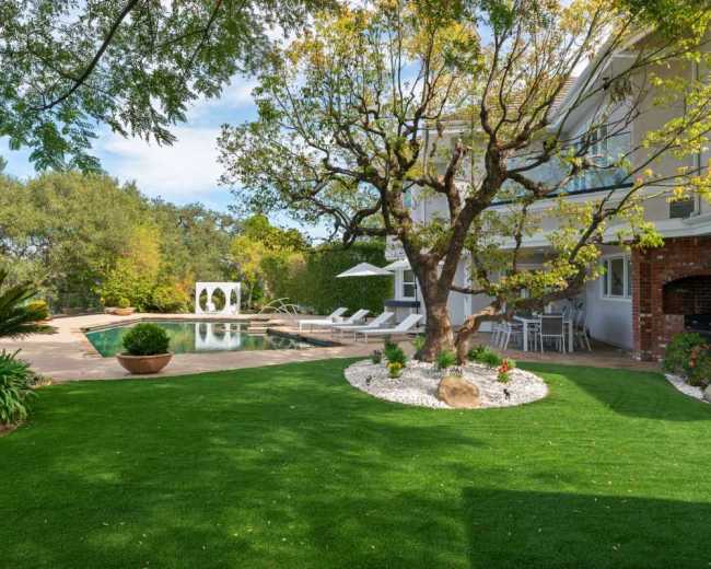The image shows a landscaped backyard featuring a swimming pool, lounge chairs, a patio area, and a large tree surrounded by greenery.