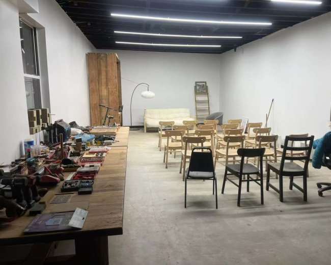 The image shows a spacious, empty room with a row of wooden chairs set up facing a wall, and a long table cluttered with various tools and materials on one side.