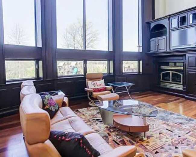 The image shows a modern living room with large windows, a cream-colored sectional sofa, a glass coffee table, and a dark wooden entertainment unit.