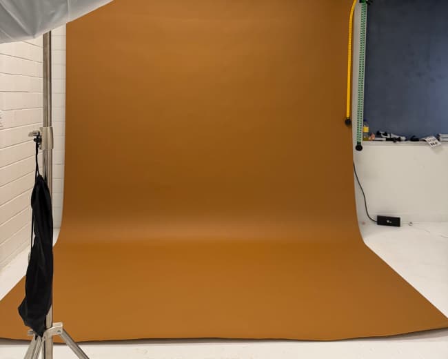 The image shows a photography studio setup with a large brown backdrop and a light source positioned overhead.