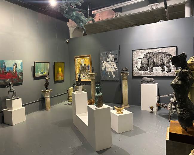 The image depicts an art gallery displaying various sculptures and paintings on gray walls, with artworks arranged on pedestals and hung at eye level.