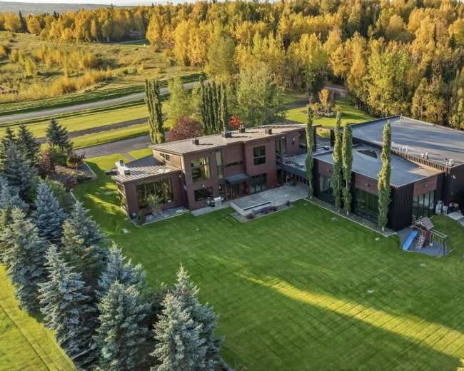 The image shows a large modern house surrounded by well-manicured lawns and lined with trees, set against a backdrop of autumn foliage.