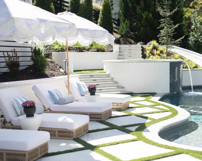 The image shows a luxury outdoor pool area with white lounge chairs, an umbrella, and a landscaped pathway leading to steps and a water feature.