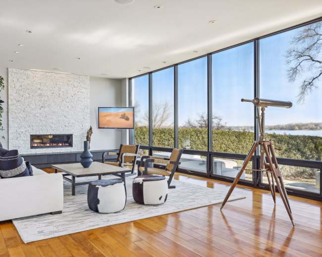 A modern living room features large windows with a view of the outdoors, a sleek fireplace, and contemporary furniture, including a telescope.
