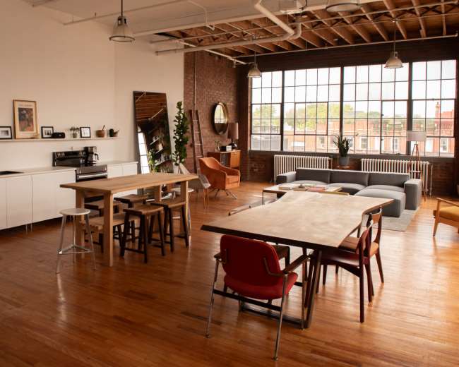The image shows a spacious, modern loft with wooden floors, featuring a kitchen area, a dining table, seating arrangements, and large windows allowing natural light to enter.