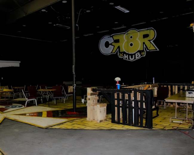 The image shows a dimly lit room with tables and chairs arranged in a creative space, featuring a painted floor design and a wall displaying the text "CR8R HUB."