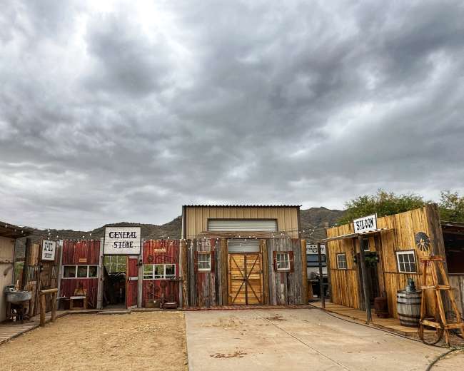A rustic facade featuring several weathered wooden structures labeled "General Store" and "Saloon" under a cloudy sky.