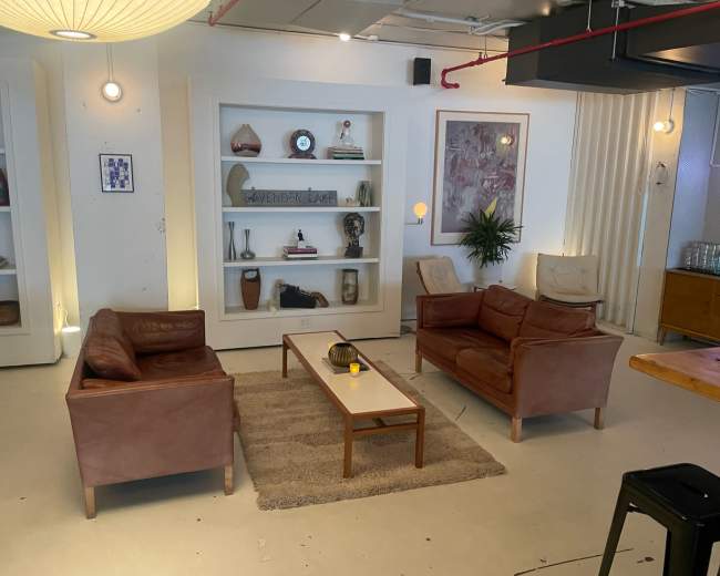 The image shows a cozy lounge area with two leather sofas facing a central coffee table, surrounded by decorative shelves and a large artwork on the wall.